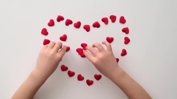 Καρδιά από γυαλιστερές κόκκινες μικρές διακοσμητικές καρδιές σε λευκό φόντο με παιδικά χέρια — Αρχείο Βίντεο