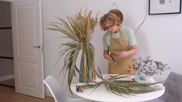 Portrait of senior female in yellow apron takes photos of dried flowers and herbs — Stock Video