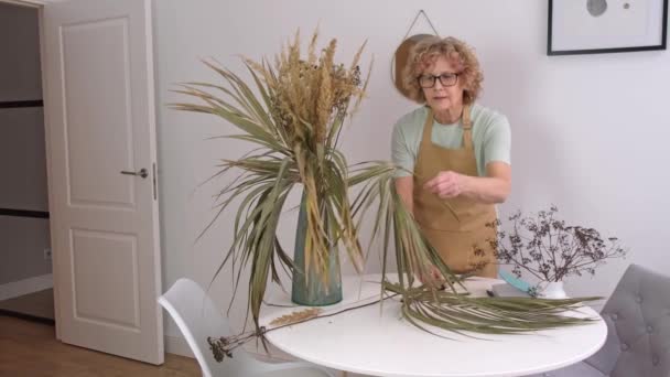 Porträtt av senior hona i gult förkläde arrangerar torkade blommor och örter — Stockvideo