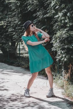 Turkuaz elbiseli ve şapkalı bir kadın parktaki ara sokakta poz veriyor, parmakları içe dönük duruyor..