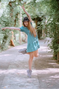 Uzun saçlı, kısa elbiseli ve şapkalı bir kadın parkta poz veriyor. Ayak parmağının üzerinde duruyor ve ellerini kaldırıyor.