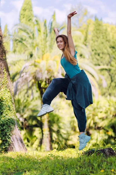 Fille en turquoise vêtements hip-hop gelé dans un saut dans une position des mains vers le haut et lever le genou dans un parc vert d'été par une journée ensoleillée — Photo