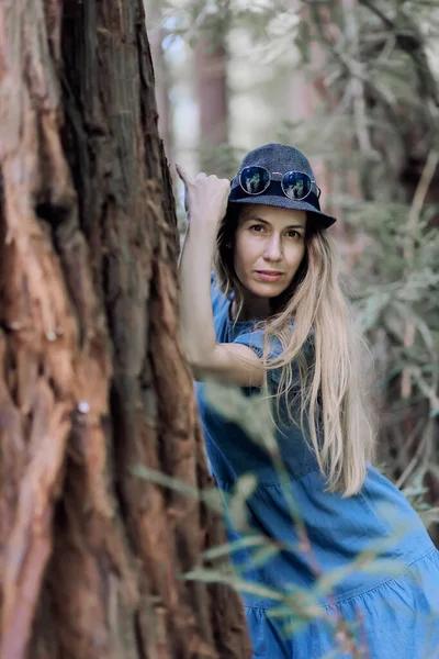 Uma menina de cabelos longos em um chapéu está com a mão apoiada contra uma árvore no parque. — Fotografia de Stock