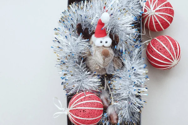 一只长着鼓鼓的眼睛的有趣的小玩具鹿躺在镶着红色圣诞球的银光里。圣诞装饰. — 图库照片