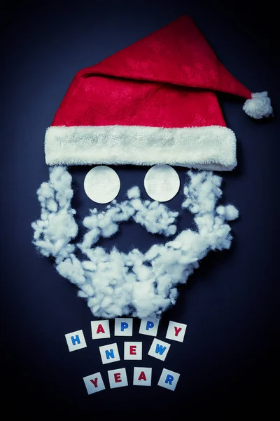 Ein roter Hut mit fasergefülltem Schnurrbart und Bart und Baumwollpolstern für die Augen auf dunkelblauem Hintergrund. Lustiges Santas-Gesicht. Neujahrskarte — Stockfoto