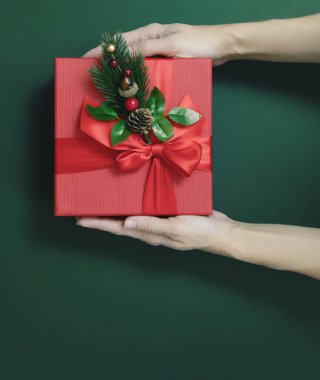 Eller, yeşil arka planda dekoratif bir köknar filizi olan kare kırmızı bir hediye kutusu tutuyor. Üst Manzara. Tatil hediyesi.