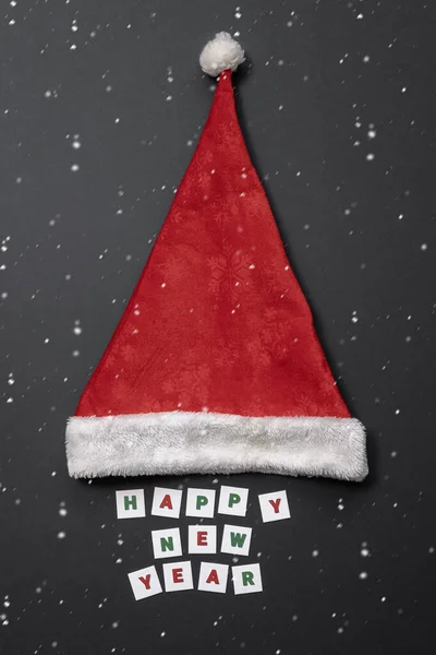 Weiße Schneekörner fliegen vor dem Hintergrund der Inschrift Frohes Neues Jahr und einer roten Weihnachtsmütze auf dunkelgrauem Hintergrund. — Stockfoto