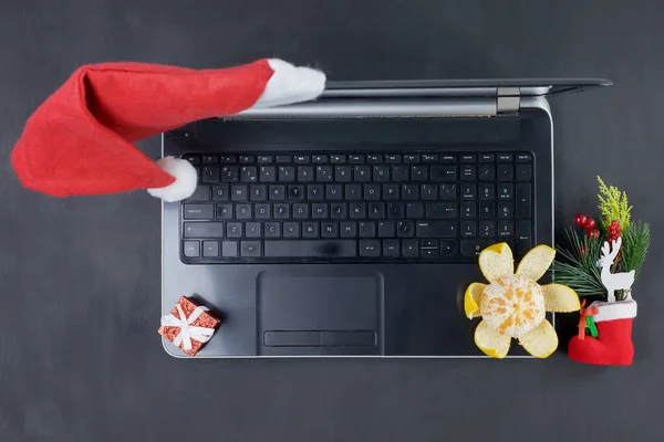 Otwarty laptop stoi na szarym tle, nosząc czapkę Mikołaja, małe pudełko, obraną mandarynkę i ozdobną zabawkę świąteczną. Układ graficzny — Zdjęcie stockowe