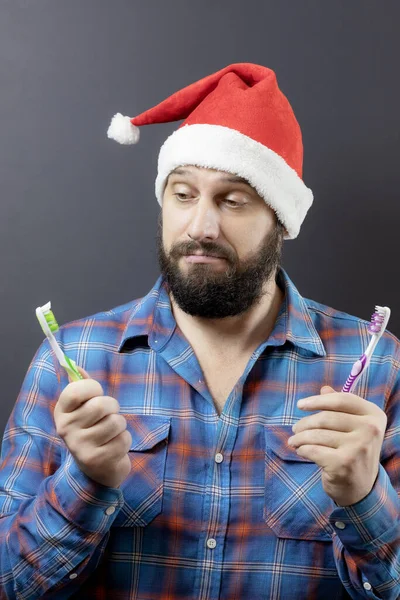 A pensive bearded man in a Santa hat is choosing from two toothbrushes. Preparing for Christmas. Fake santa — Stock Photo, Image