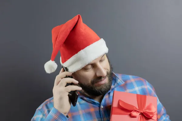 A man in a santa hat with a red box with a gift talking on a cell phone — Stock Photo, Image