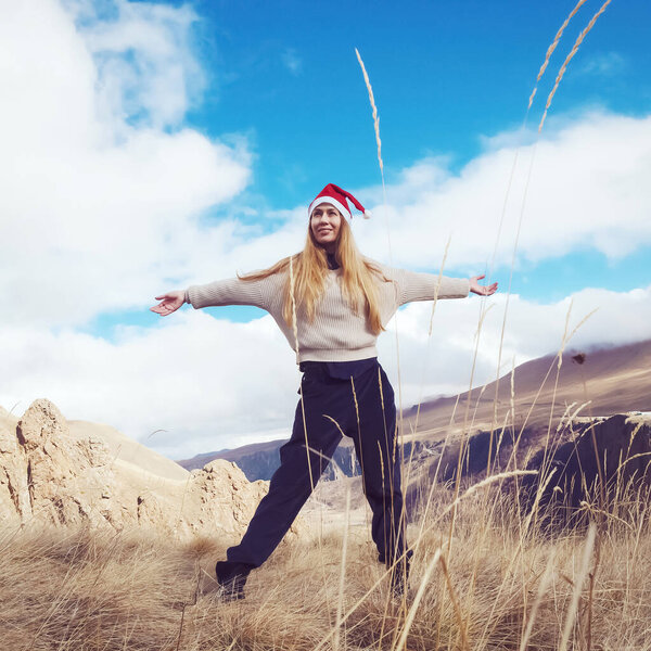 A woman in a santa claus hat stands against the background of clouds and mountains spreading her arms to the sides, rejoicing in nature.