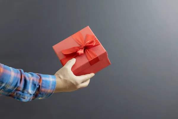 De hand van een man in een geruit shirt houdt een rode doos met een feestelijk geschenk op een grijze achtergrond — Stockfoto