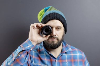 Örgü şapkalı ve ekose gömlekli sakallı bir adam yuvarlak bir lensten bakar.