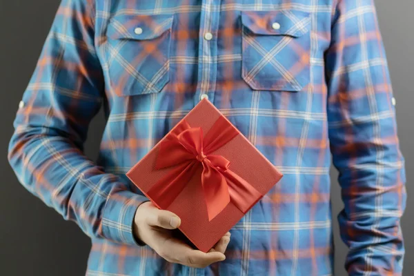 Een rode geschenkdoos met een strik in de hand van een man in een blauw geruit shirt. Feestelijke verrassing — Stockfoto