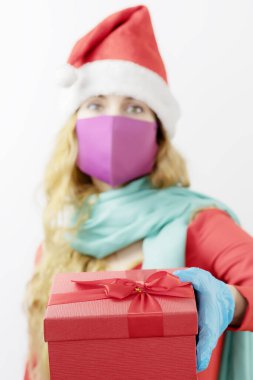 Kızların elinde Noel hediyesi olan bir kutu. Tıbbi maske ve eldiven takmış Noel Baba şapkalı bir kadın. Noel için tam koruma. Steril hediye. Hediyeye odaklan.