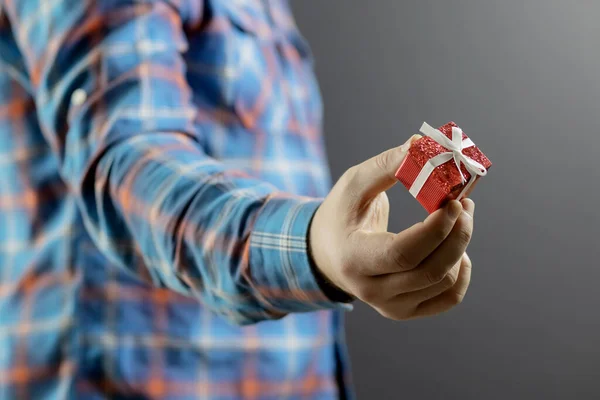 Een kleine rode doos met een cadeautje in de hand van een man in een blauw geruit shirt. Een kleine verrassing. Concentreer je op de hand. Geschenkvervaging — Stockfoto