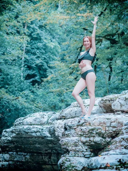 Een glimlachende vrouw in een bikini staat met haar hand opgeheven aan de rand van een kloof omringd door loofbos. Buitenactiviteiten — Stockfoto