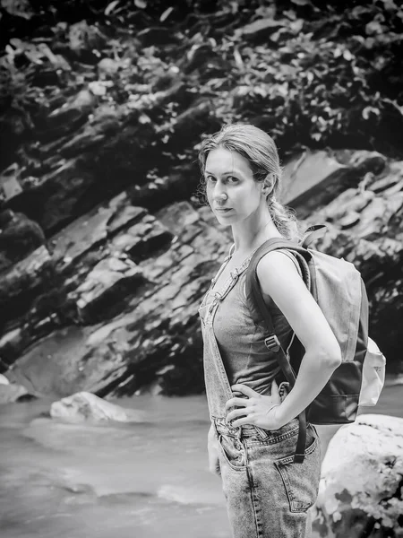 Ένα κορίτσι με τζιν φόρμα με ένα σακίδιο στην πλάτη στέκεται στην όχθη του ποταμού με φόντο ένα βράχο. Πεζοπορία. Ασπρόμαυρη φωτογραφία — Φωτογραφία Αρχείου
