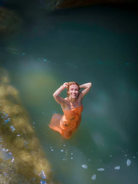 Ένα κορίτσι με ένα βρεγμένο φόρεμα χαμογελώντας κοιτάζει ψηλά ενώ στέκεται στο νερό σε ένα φαράγγι φαράγγι φωτισμένο από τον ήλιο — Φωτογραφία Αρχείου