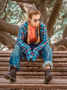 Ekose gömlekli, uzun kollu, büyük beden kotlu ve kaba ayakkabılı bir kız parkta bir bankta oturuyor..