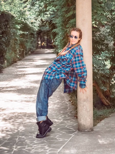 Kobieta z warkoczami w luźnym, przerośniętym stroju stoi zgięta w dolnej części pleców, opierając się o kolumnę w parku przy rozwidleniu na drodze.. — Zdjęcie stockowe