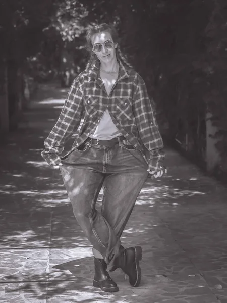 Una ragazza con le treccine in camicia scozzese e jeans molto larghi si trova in una posa curtsy in un vicolo in un parco. Foto in bianco e nero — Foto Stock