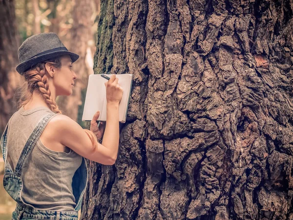 Une jeune fille aux cheveux blonds dans un chapeau prend des notes dans un cahier appuyé sur un tronc massif d'un arbre forestier — Photo