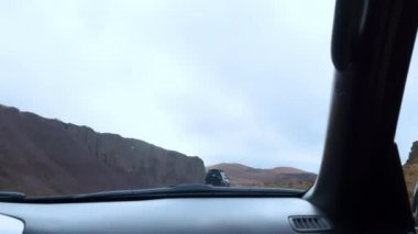Çamurlu ve engebeli bir yolda Jeep turu. Ön koltuktan ön cama ateş ediyor. Arabalar önümüzde ve büyük kayalar görünüyor..