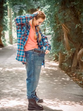Ekose gömlekli ve büyük beden kotlu bir kadın, parkın beton yolunda pantolonunun içine korkmuş bir şekilde bakıyor..