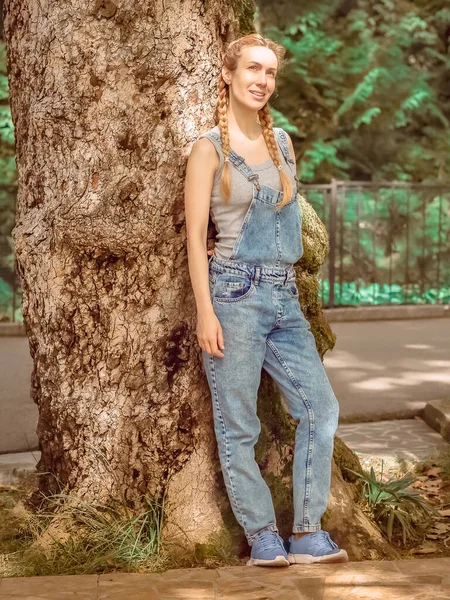 Χαμογελαστή κυρία με κοτσίδες σε denim φόρμες στέκεται ακουμπισμένη στο πορτ-μπαγκάζ ενός τεράστιου δέντρου στο πάρκο μια καλοκαιρινή μέρα. Συγκεντρώσου στα μάτια. — Φωτογραφία Αρχείου