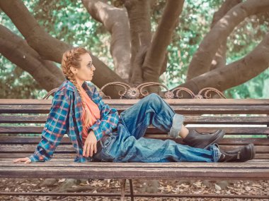 Ekose gömlekli at kuyruklu kadın, büyük beden kot pantolon ve sert botlar parkın gölgeli bölgesinde bir bankta oturuyor.