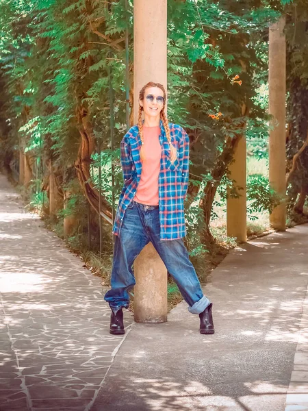 Улыбающаяся девушка с косичками в клетчатых длинных джинсах и грубых сапогах стоит у вилки в парке, опираясь на колонну — стоковое фото