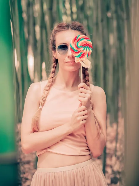 Dame met vlechtjes bedekt een oog met een veelkleurige ronde lolly terwijl ze in een groene bamboe bos staat — Stockfoto