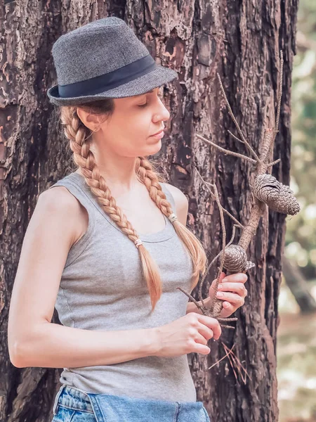 Ένα ξανθό κορίτσι με καπέλο κρατά ένα κλαδί με κώνους ενώ στέκεται κάτω από ένα δέντρο στο δάσος. — Φωτογραφία Αρχείου