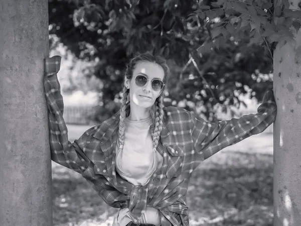 Μια χαριτωμένη κυρία με γυαλιά ηλίου με δύο πλεξούδες σε καρό πουκάμισο με πολύ μακριά μανίκια στέκεται ανάμεσα στις κολώνες του πάρκου. Ασπρόμαυρη φωτογραφία — Φωτογραφία Αρχείου