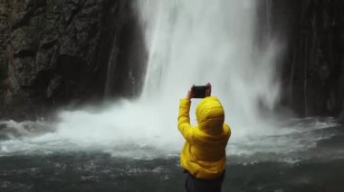 Sarı ceketli bir kız, suyun ters hareketinde büyük bir şelalenin telefonunda video çekiyor. Video düzenleme. Turizm ve eğlence konsepti