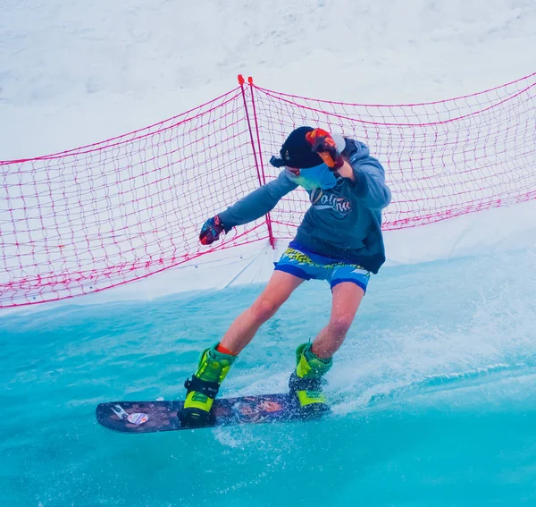 Russland, Sotschi 11.05.2019. Ein Mann in kurzen Hosen mit einer Go-Pro-Kamera auf dem Kopf überquert auf einem Snowboard einen Wasserpool. Snowboard-Wettbewerb auf dem Wasser im Skigebiet Krasnaja Poljana. — Stockfoto