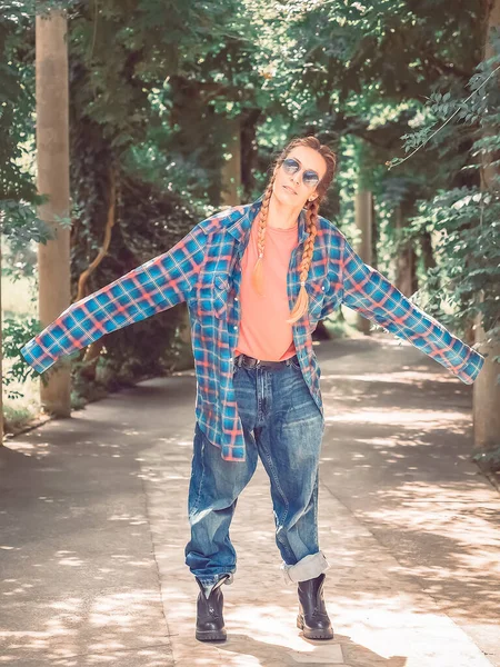 A girl with two braids in a plaid shirt with overly long sleeves and huge jeans stands spreading her arms to the side in the park on a green alley — стоковое фото