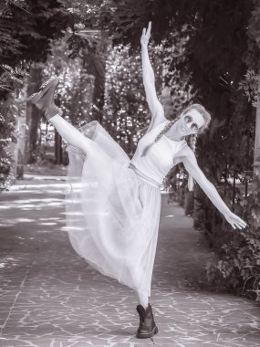 Balerin etekli ve sert çizmeli bir kız, kollarını parktaki bir ara sokakta sonuna kadar açıp sallıyor. Siyah beyaz fotoğraf