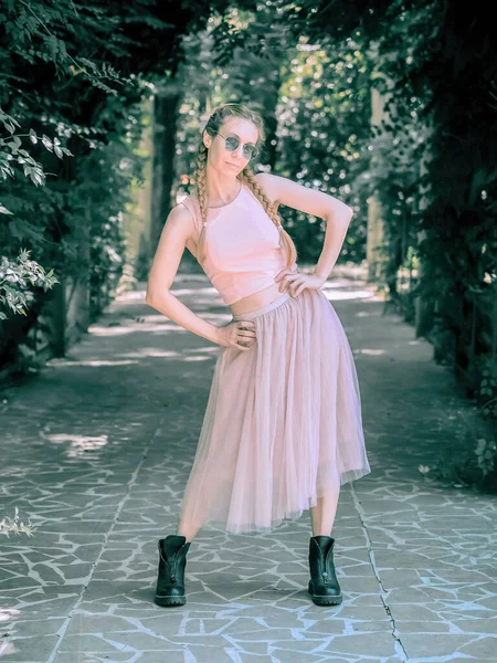 A lady with pigtails in a tutu and rough boots stands with her hands on her belt and legs apart in an alley in a summer park. Post-processing. Artistic effect. — Fotografia de Stock