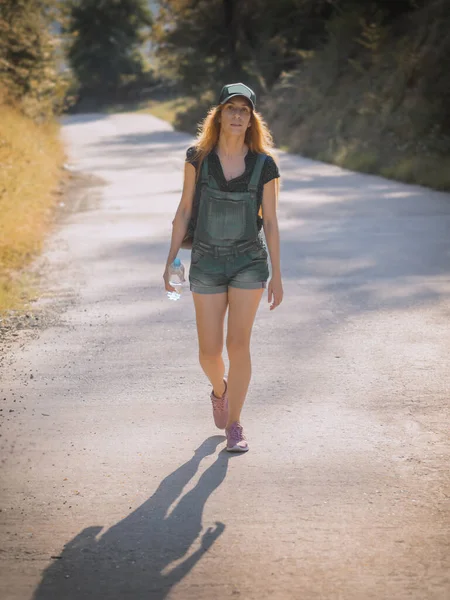 Ένα κορίτσι με κοντά denim φόρμες και ένα καπέλο περπατά κατά μήκος ενός άδειου δρόμου σε μια ηλιόλουστη καλοκαιρινή μέρα — Φωτογραφία Αρχείου