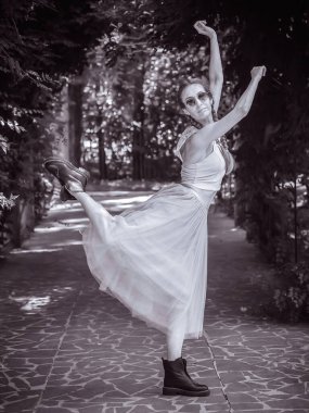 At kuyruklu, etekli ve çizmeli bir kadın yaz parkındaki dar bir sokağa geri döner. Çağdaş bale. Siyah beyaz fotoğraf. Bulanık odak