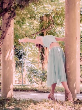 Bale eteği ve topuklu ayakkabılı bir kız gölgeli bir yaz parkının sütunlarının yanında sırtüstü duruyor. Bulanık odak