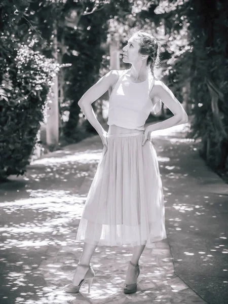 Uma menina com tranças em uma saia de tule leve e sapatos de salto alto fica em uma pose em um beco no parque. Foto em preto e branco. Foco no Borrão. — Fotografia de Stock
