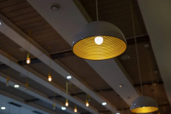 Una pantalla con una lámpara brillante cuelga en el techo de la cafetería contra el fondo de otras bombillas. Iluminación de techo en un estilo moderno —  Fotos de Stock