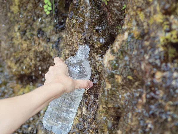 Ženská ruka naplní plastovou láhev pramenitou vodou z pramene stékajícího po skále. Jaro slz pro dívky — Stock fotografie