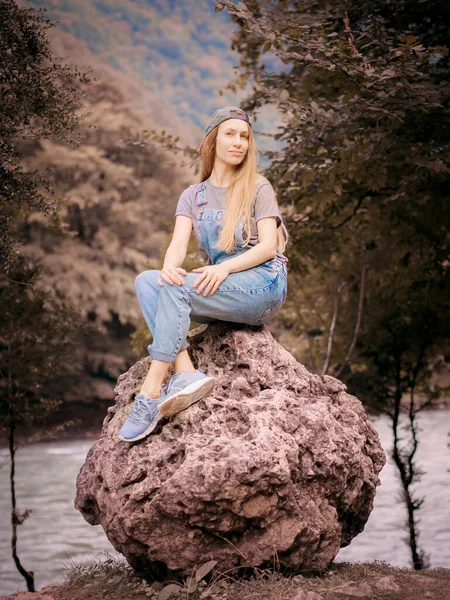 Χαμογελώντας ξανθά μαλλιά γυναίκα σε denim φόρμες κάθεται σε μια στρογγυλή μεγάλη πέτρα σε μια κοιλάδα του δάσους με φόντο ένα ποτάμι — Φωτογραφία Αρχείου
