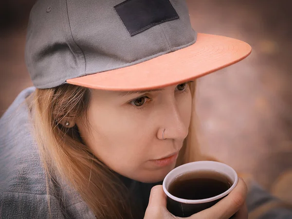 Ένα κορίτσι με καπέλο φέρνει ένα χάρτινο ποτήρι με μαύρο καφέ στα χείλη της. Κλείσιμο φωτογραφίας — Φωτογραφία Αρχείου
