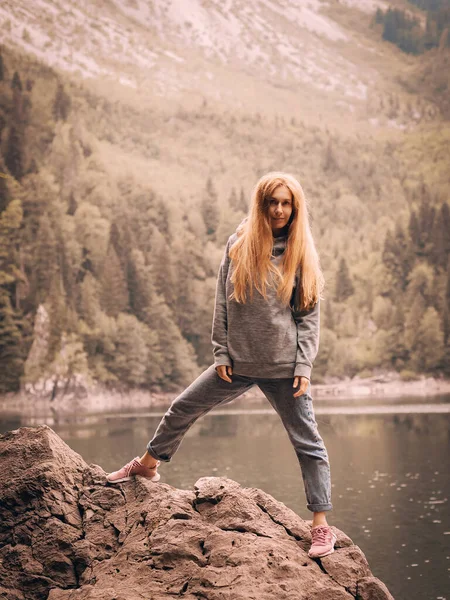 Ένας μακρυμάλλης τουρίστας στέκεται σε μια μεγάλη πέτρα με φόντο μια λίμνη σε μια κοιλάδα του βουνού — Φωτογραφία Αρχείου