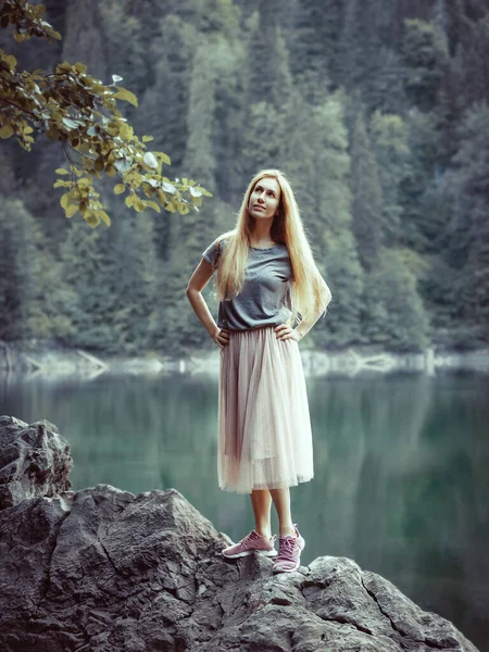 Ένα κορίτσι με μακριά φούστα στέκεται σε μια μεγάλη κοφτερή πέτρα στο φόντο μιας πεντακάθαρης λίμνης του δάσους. — Φωτογραφία Αρχείου